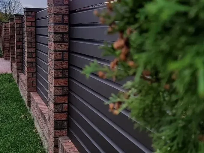 Tamsi skardinė tvora su raudonų plytų stulpais ir žaluma priekiniame plane